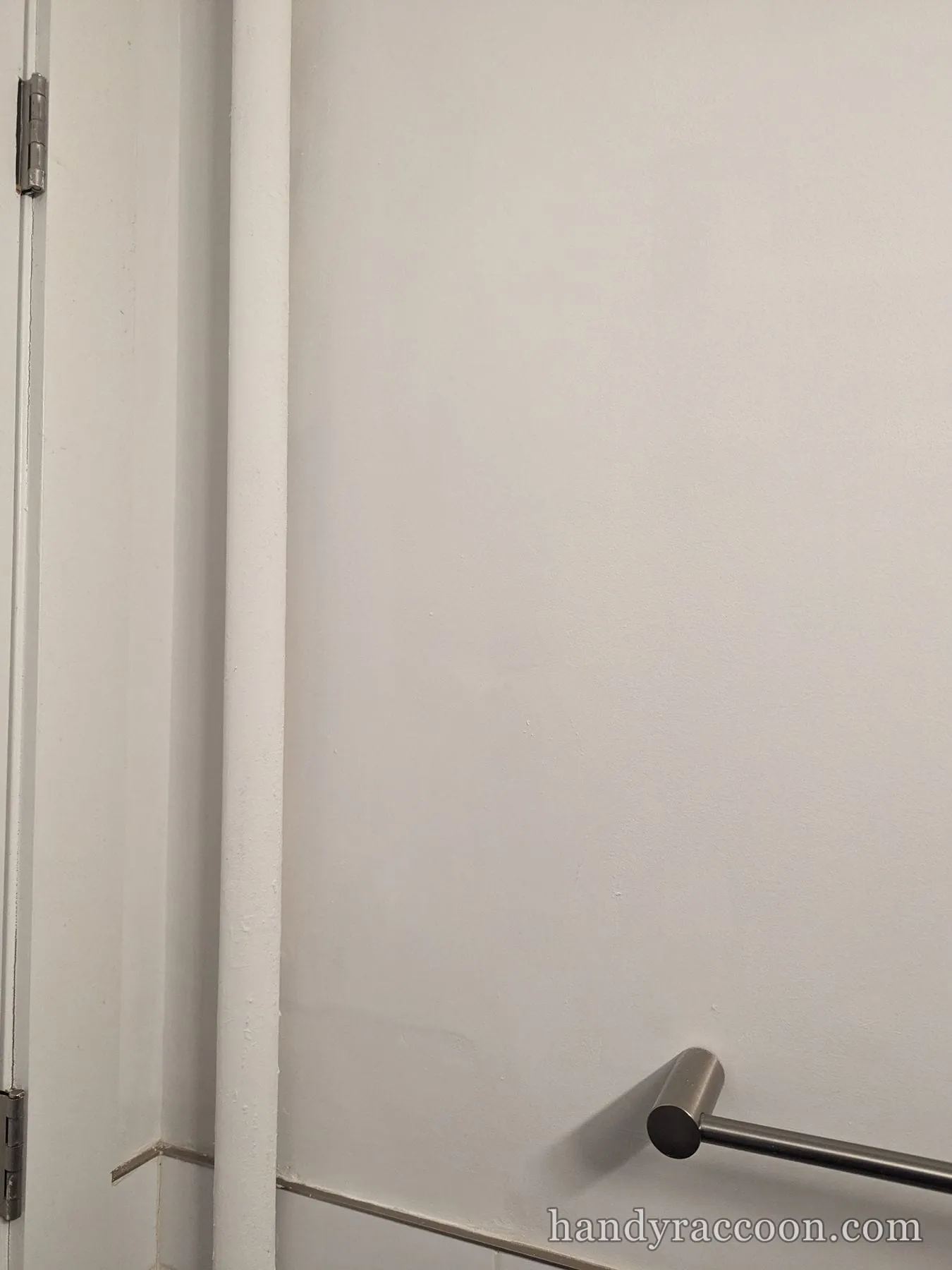 Smooth, freshly painted white bathroom wall and pipe with a door hinge and a towel bar.