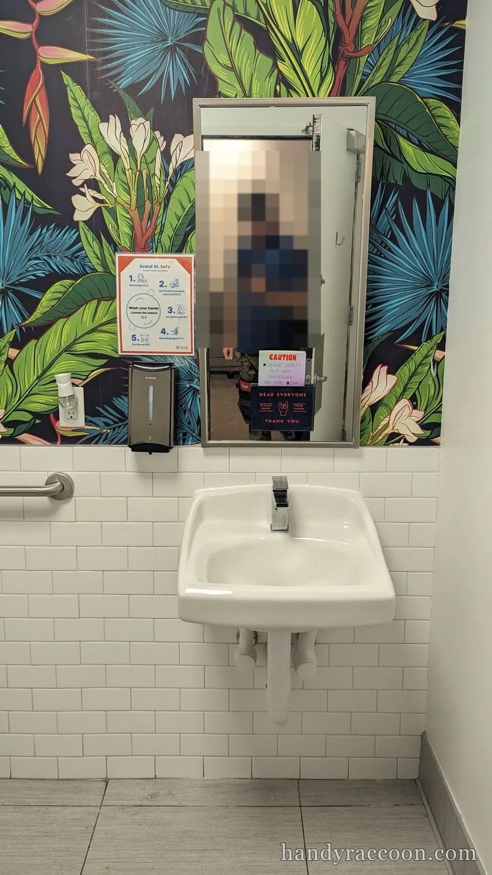 White wall-hung sink under a mirror on tiled wall with tropical wallpaper; a person is reflected in the mirror.