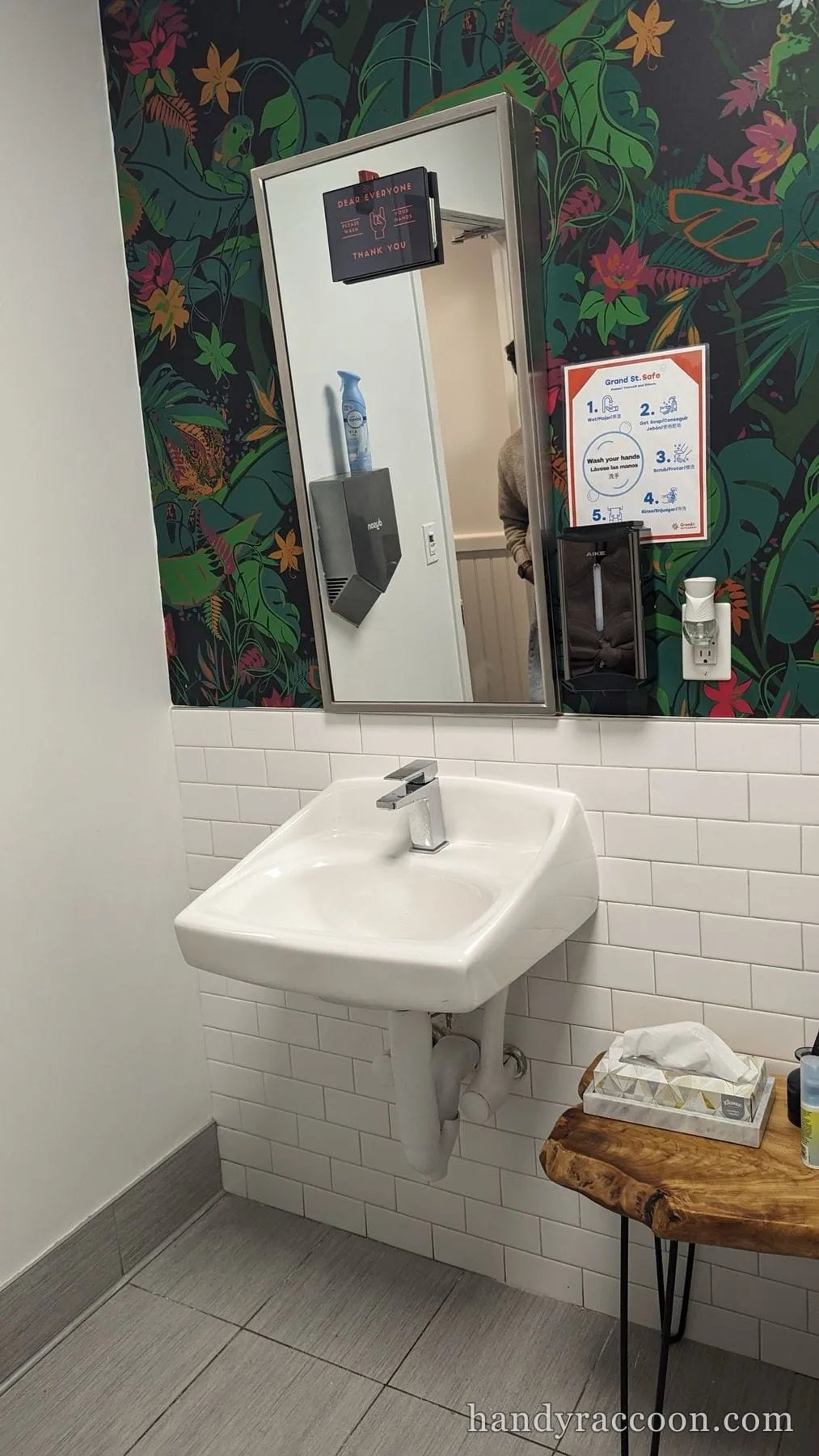 Wall-hung white sink with a square chrome faucet on white subway tile below a mirror and tropical wallpaper.