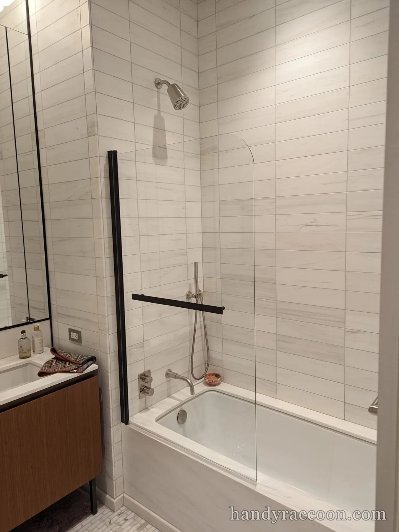 Bathtub with wall fixtures, handheld shower, and a clear glass splash panel on a marble-tiled bathroom wall.