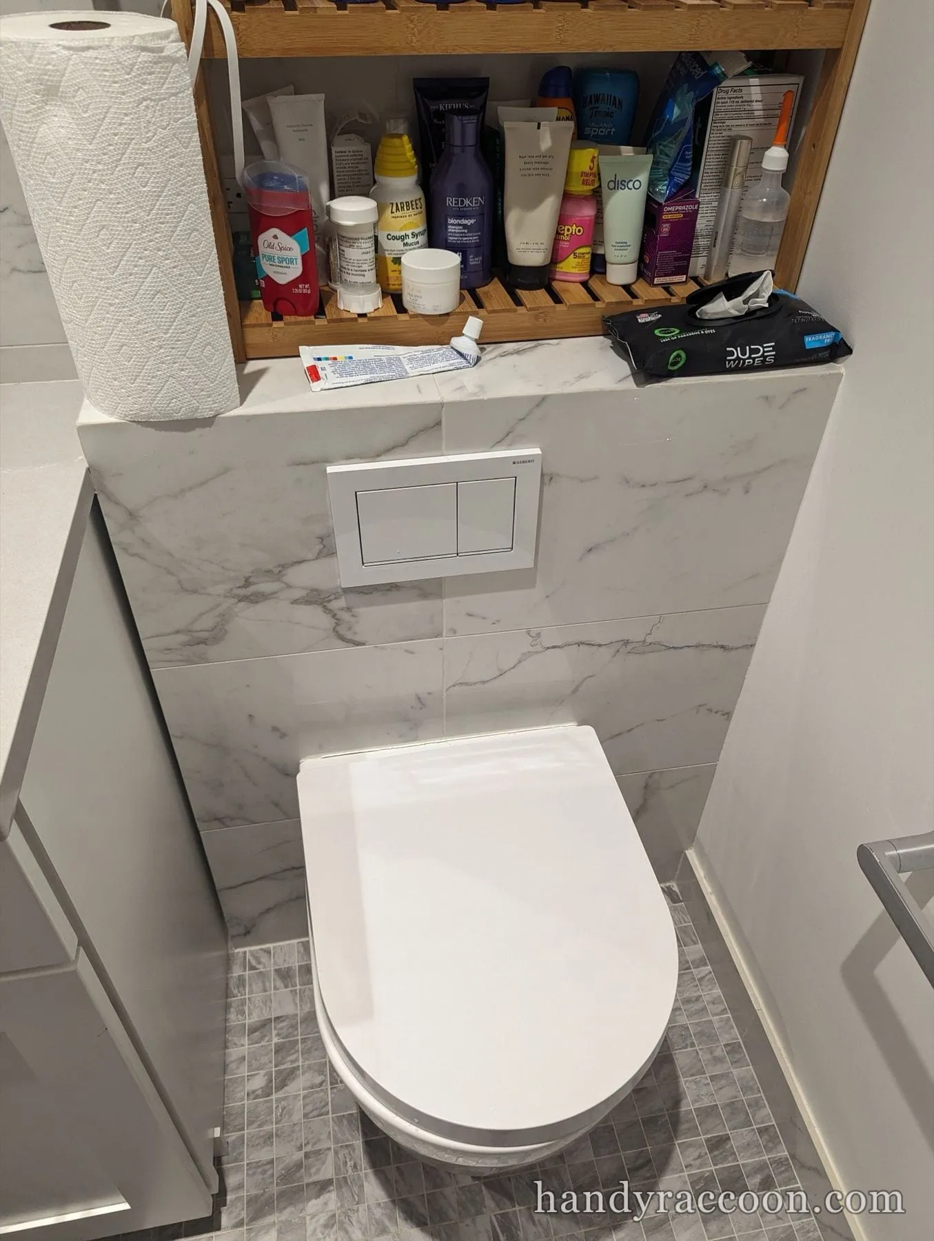 Wall-hung white toilet with a dual-flush plate on marble tile, a shelf of toiletries above.