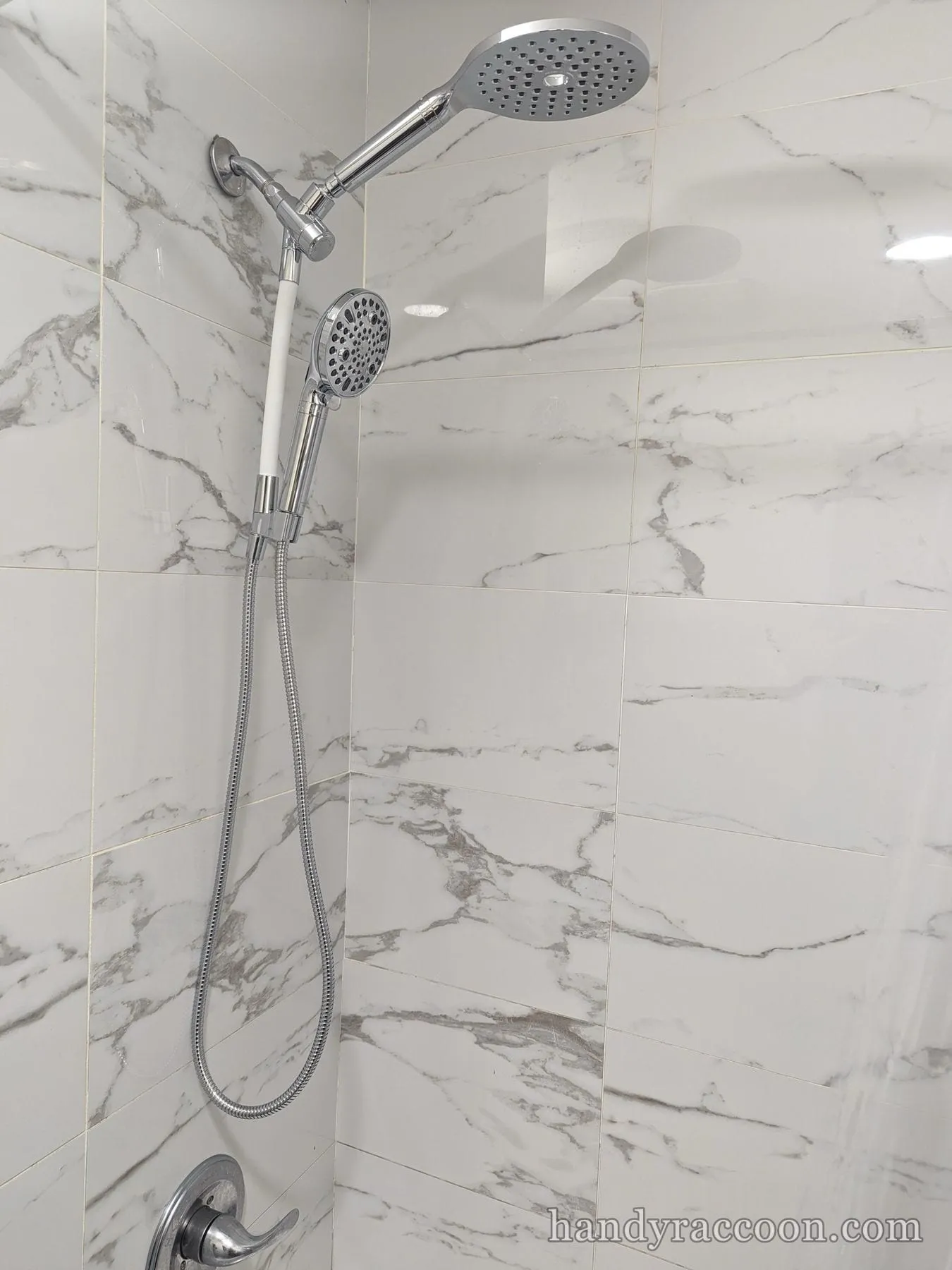 Chrome fixed shower head and handheld sprayer on a slide bar against white marble tile.