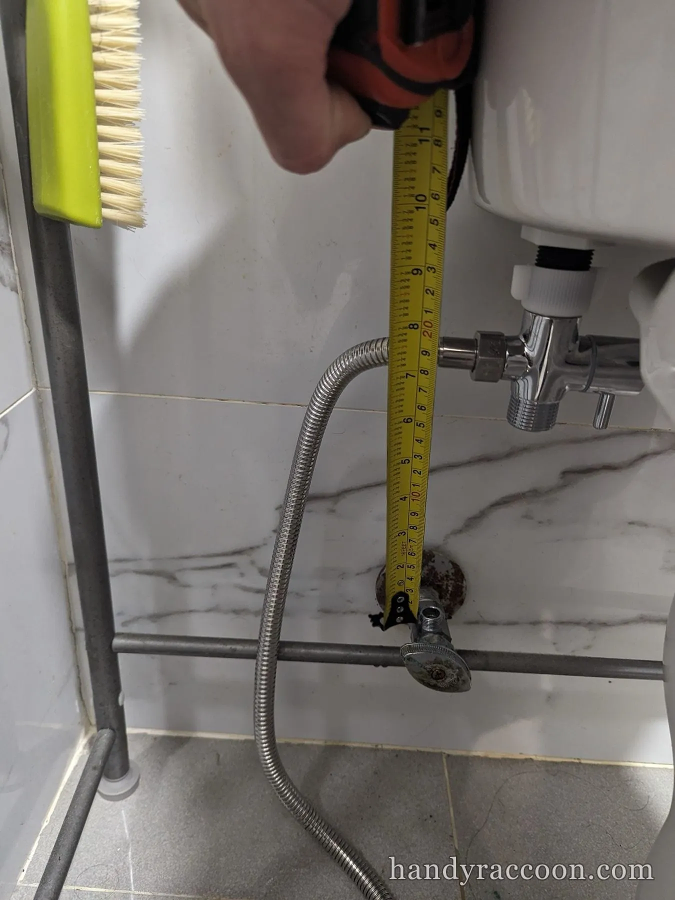 Hand holding a tape measure next to a toilet supply line and shutoff valve on marble tile.