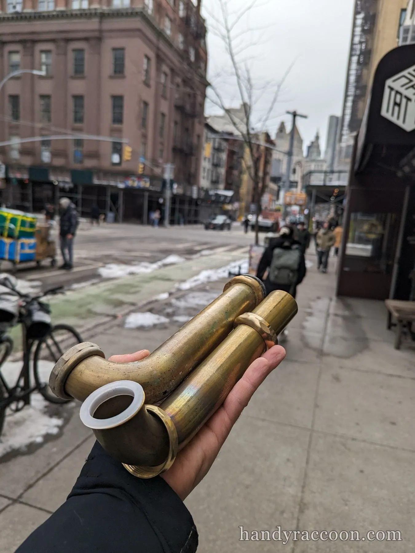 Hand holding an old brass P-trap and waste arm on a city sidewalk with buildings behind.