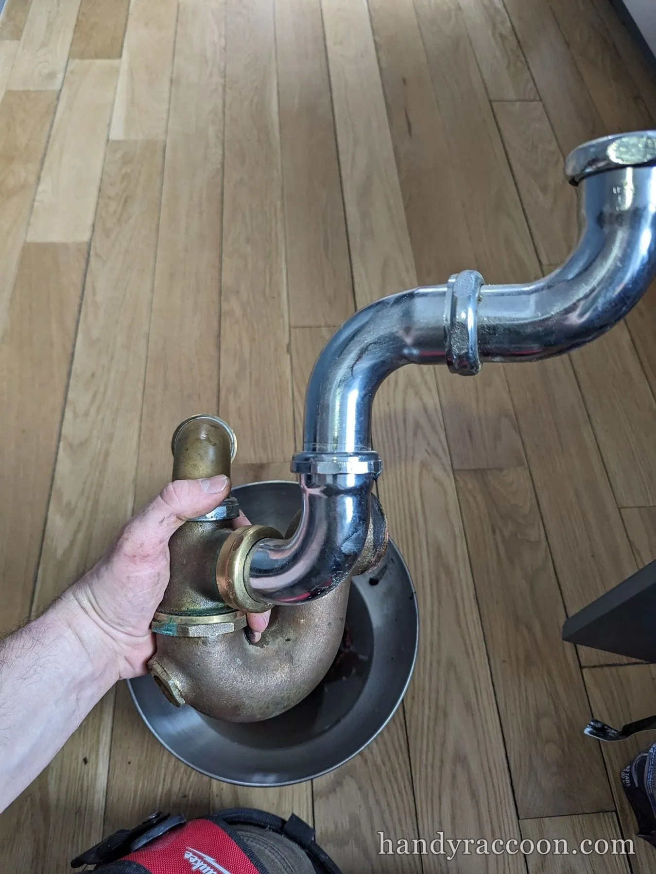 Hand holding a removed brass trap next to an installed chrome P-trap on a hardwood floor.