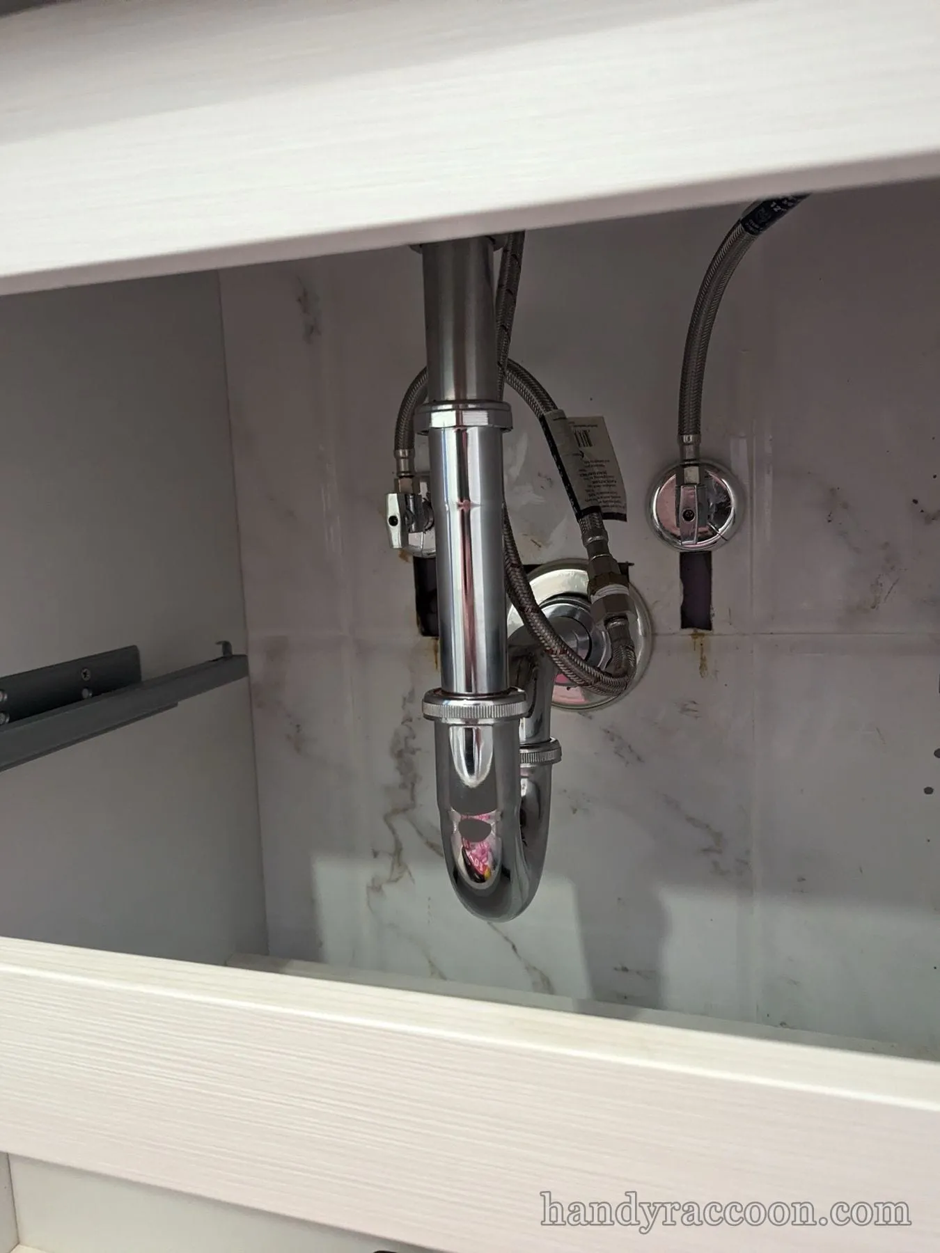 Clean chrome P-trap with braided supply lines mounted against tiled wall inside a white vanity.