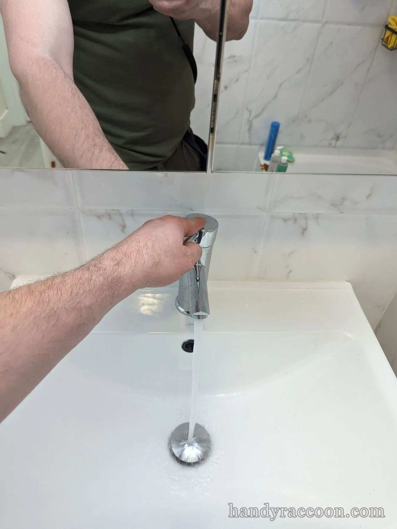 Hand operating a chrome faucet with water running into a white basin, torso reflected in the mirror.