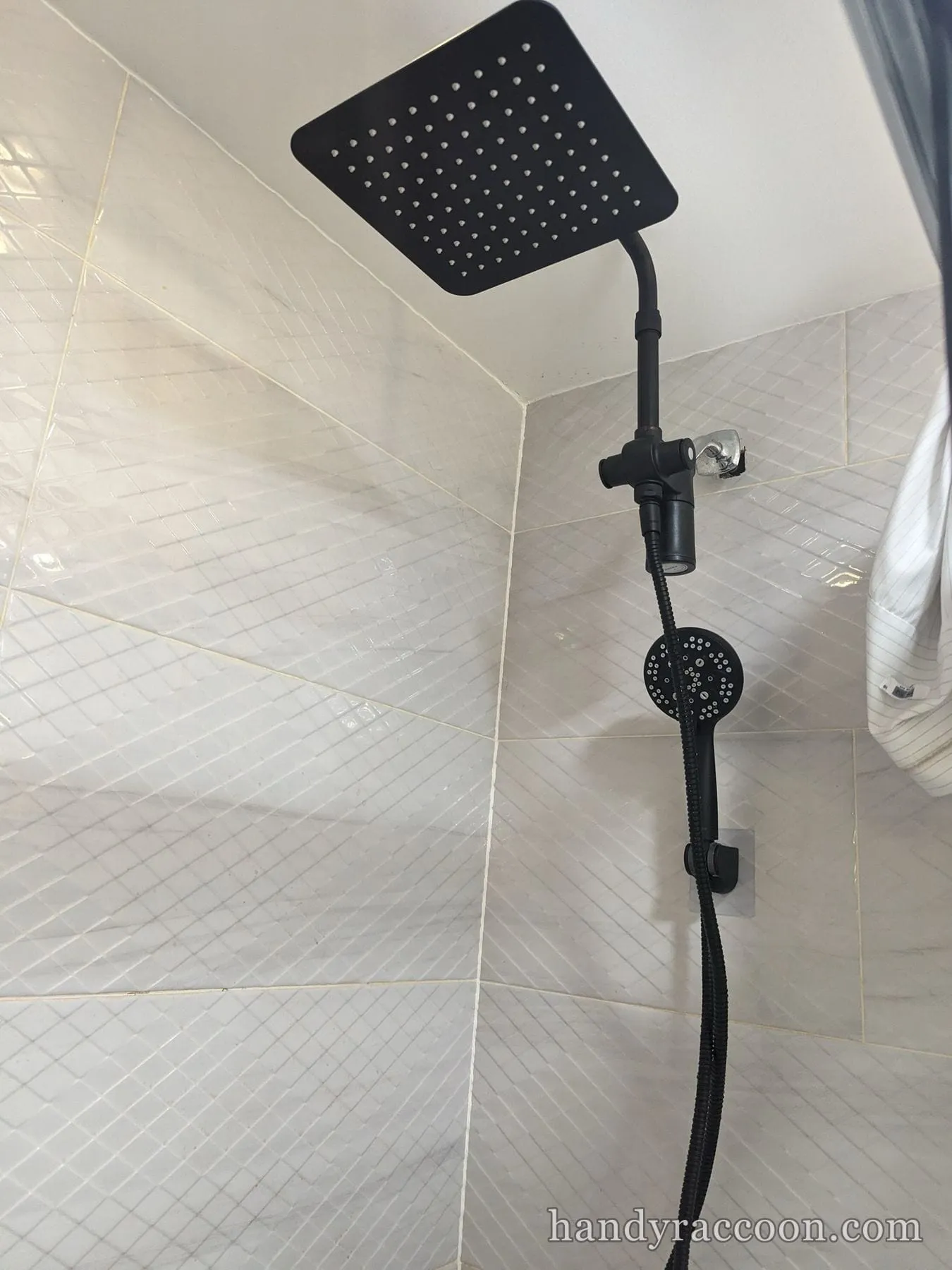 Square black rain shower head with a black handheld sprayer on hose against white diamond wall tile.