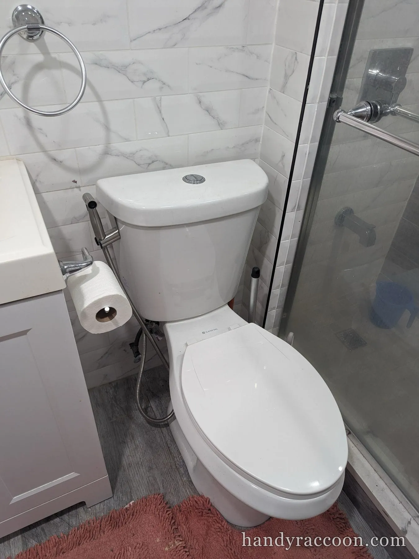 Dual-flush white toilet with a stainless handheld bidet sprayer next to a glass shower and tiled wall.