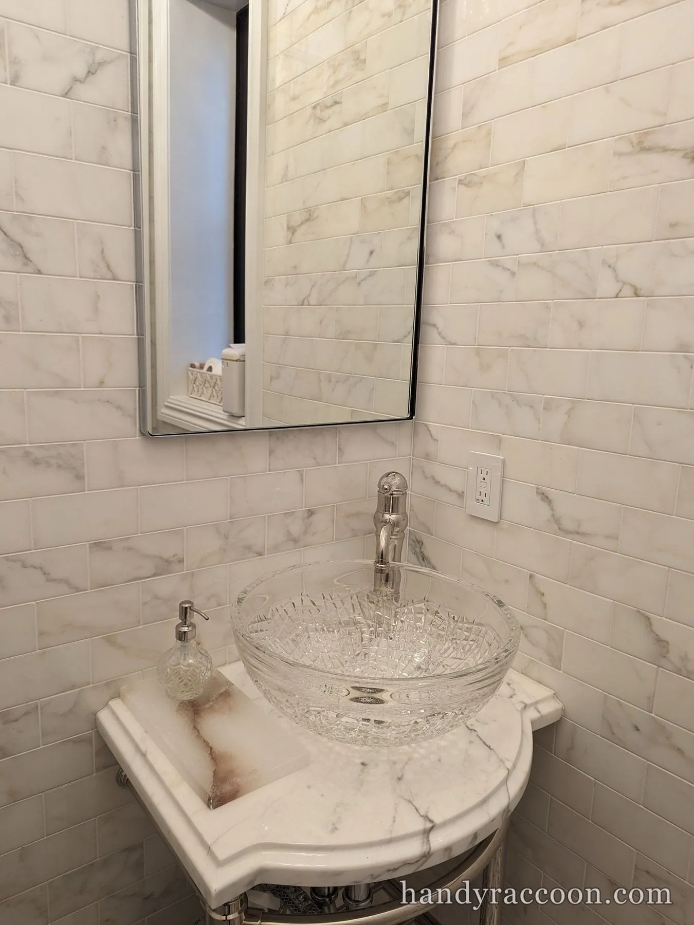 Cut-glass vessel bowl sink and chrome faucet on a light stone console against veined subway tile.