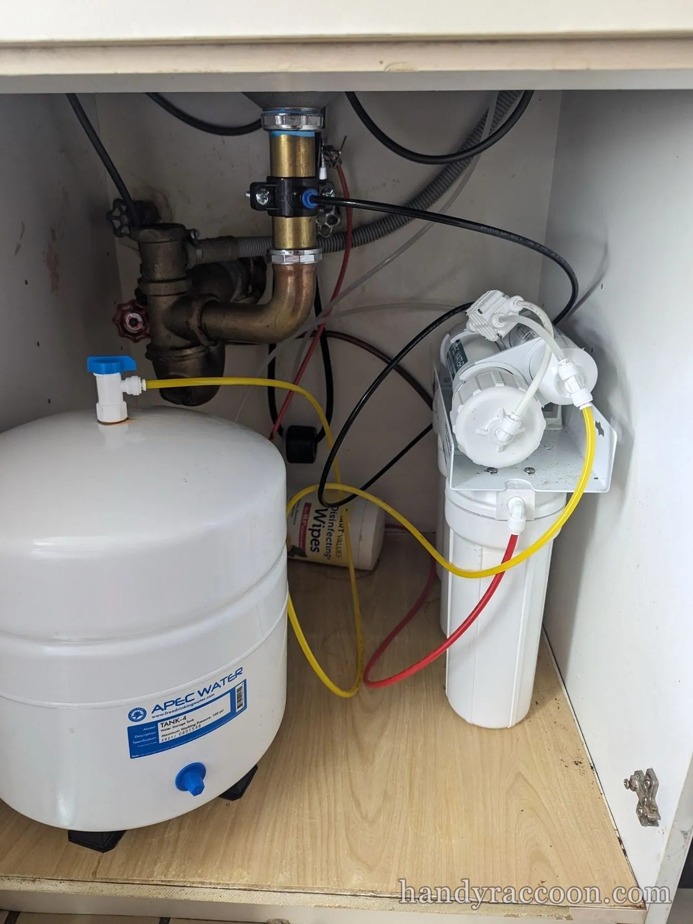 Under-sink reverse osmosis system with white storage tank, filter canister, brass P-trap and colored tubing in a cabinet.